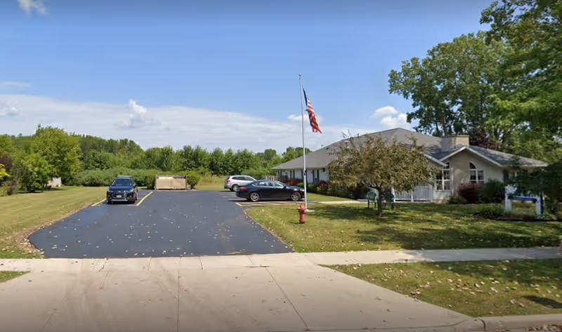 Parking lot with several cars parked next to a single-story building surrounded by trees and grass, with an American flag on a flagpole and a fire hydrant near the sidewalk.