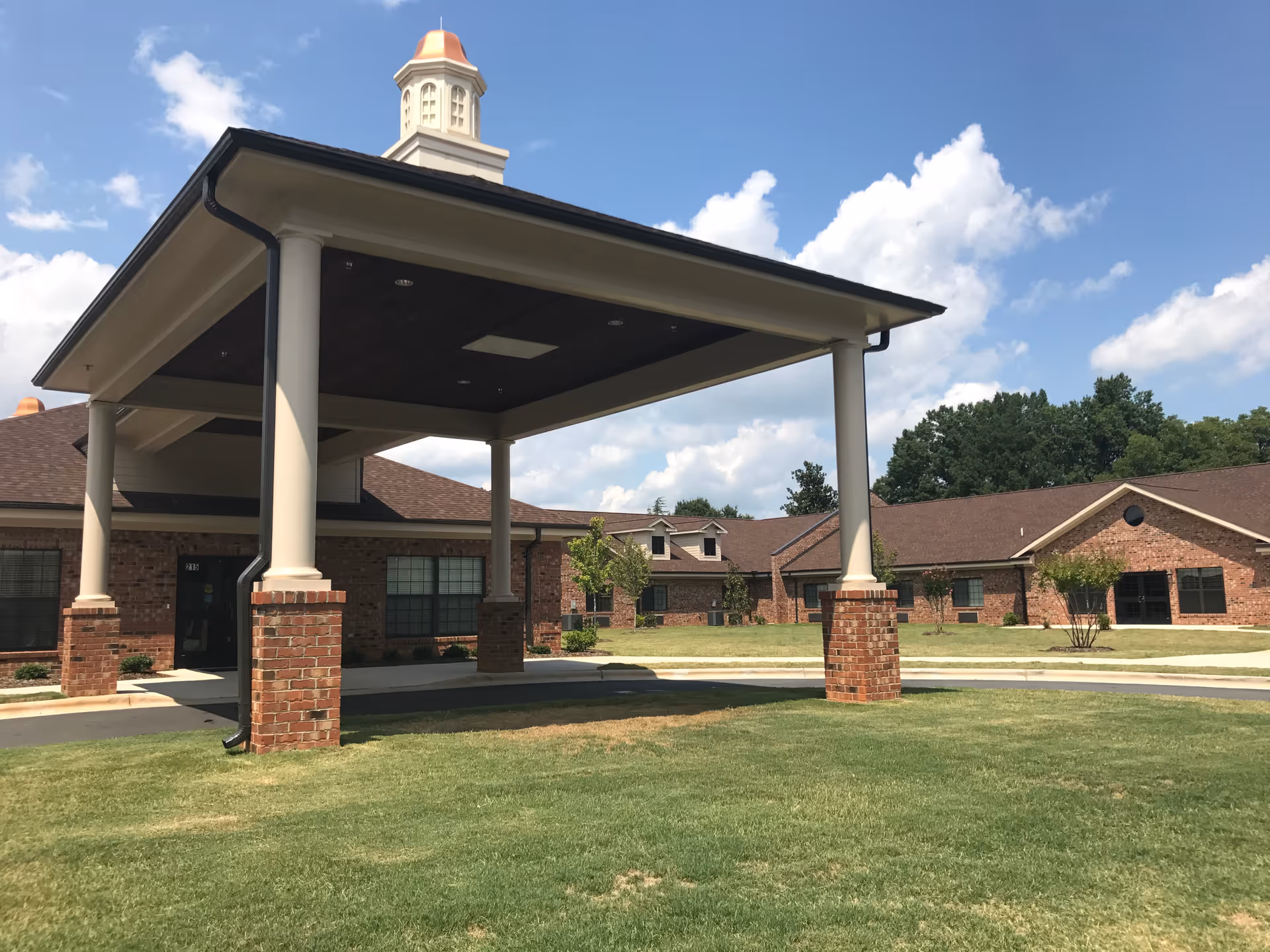 Exterior view of a single-story brick building with a covered entrance supported by white columns with brick bases. The building is surrounded by a well-maintained lawn and trees under a partly cloudy blue sky.