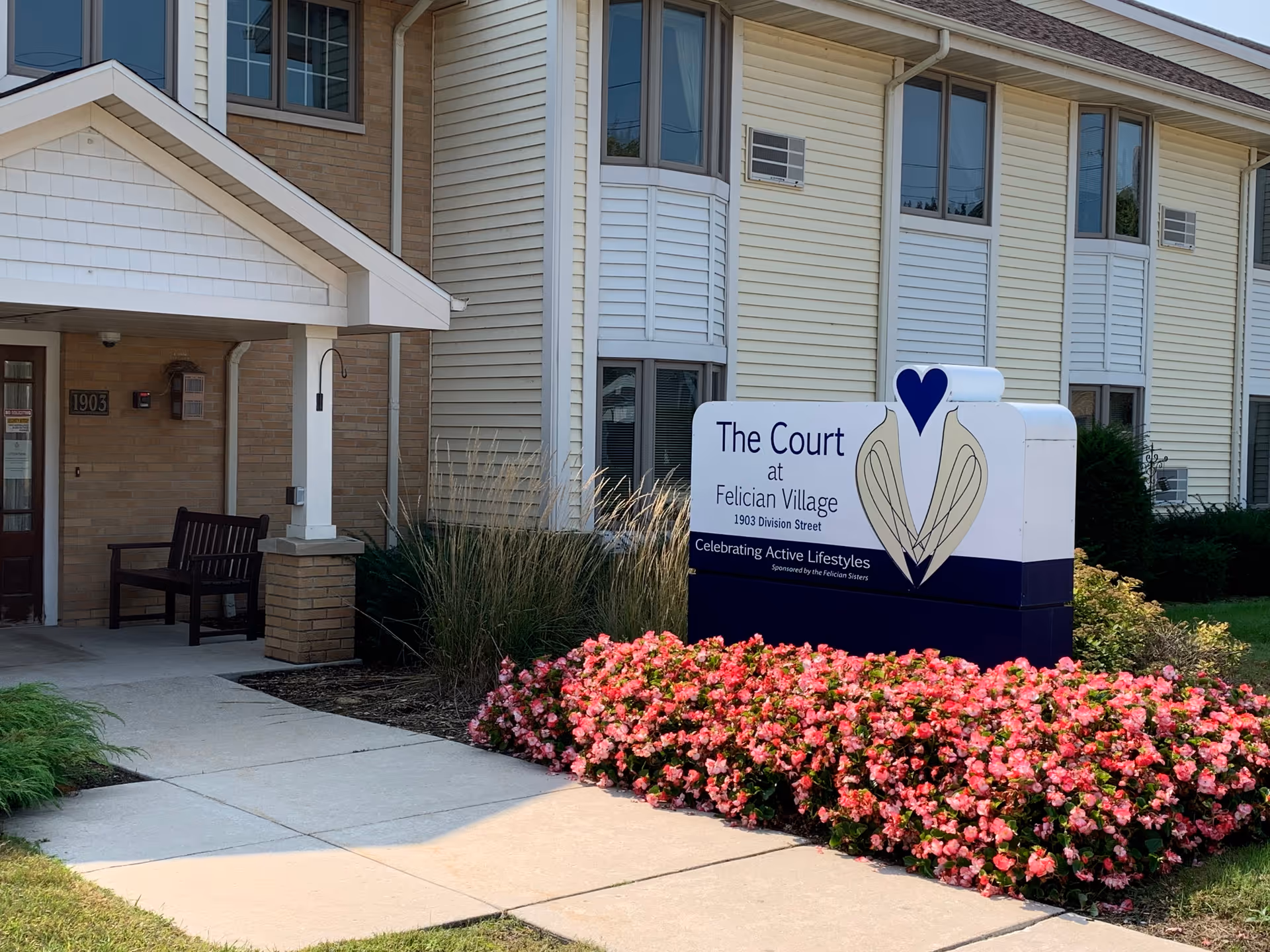 Front view of The Court at Felician Village building with a sign and pink flowers.