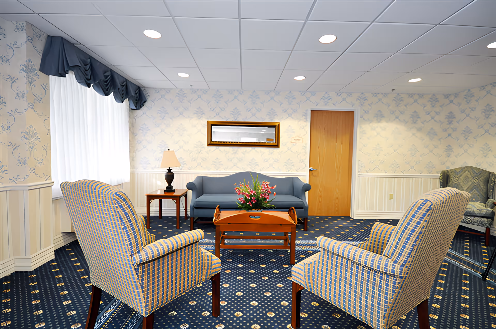 A cozy sitting area in a nursing and rehab facility featuring two patterned armchairs facing a blue sofa. A wooden coffee table with a floral arrangement is centered between the chairs and sofa. The room has blue patterned carpet, light wallpaper with a subtle design, a wooden door, a side table with a lamp, and a window with white curtains and a dark valance.