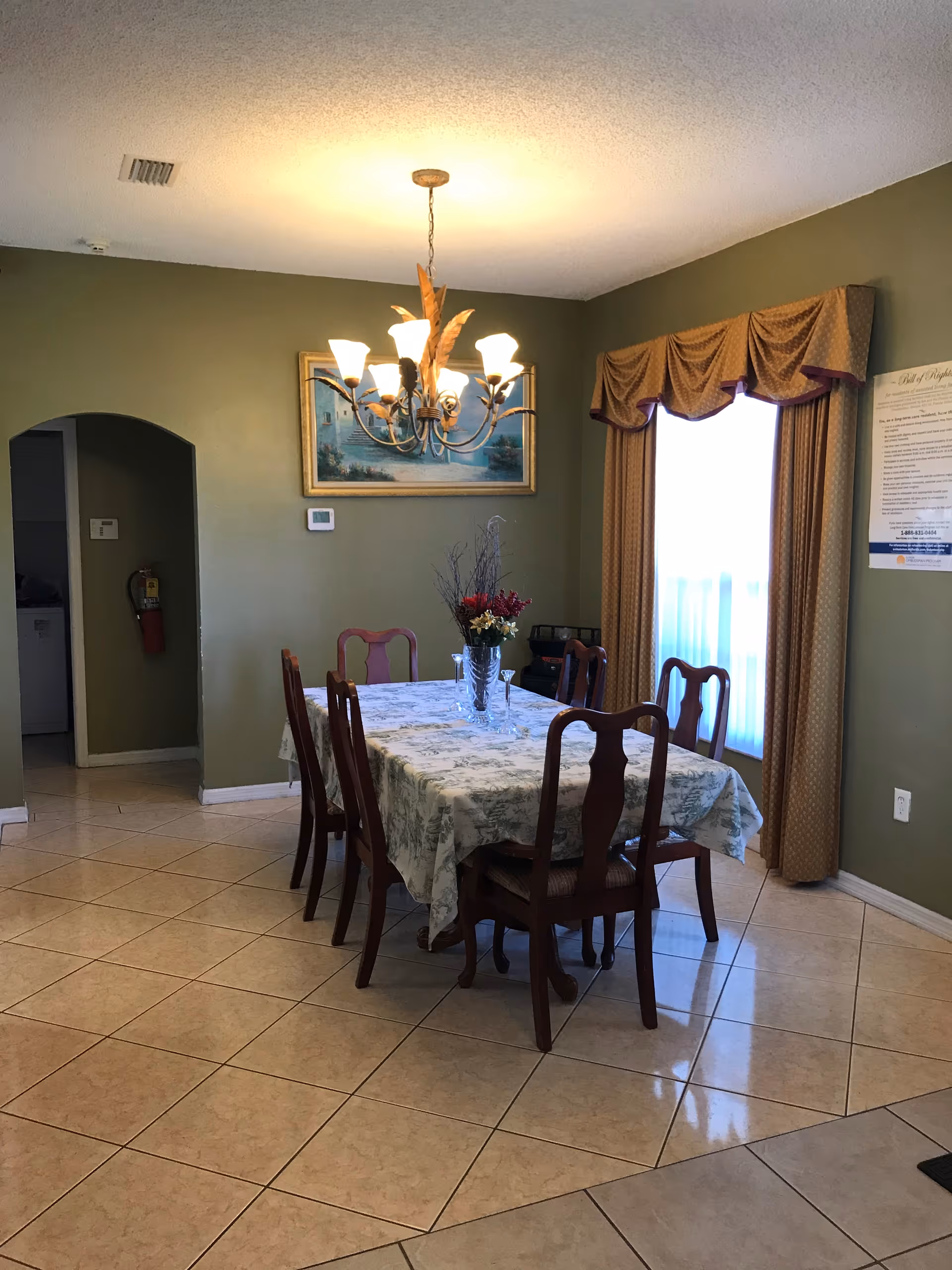 A dining room with a rectangular table covered with a floral tablecloth and six wooden chairs around it. A chandelier with multiple lights hangs above the table. There is a window with gold curtains on the right wall and a framed painting on the green wall behind the table. The floor is tiled with light-colored tiles.