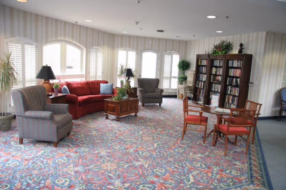 Spacious senior living lounge with red sofa, armchairs, bookshelf, and patterned carpet.
