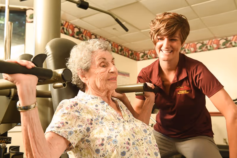 An elderly woman with curly gray hair wearing a floral blouse is using a weight machine for exercise, assisted by a smiling female staff member in a maroon shirt in a fitness or therapy room.