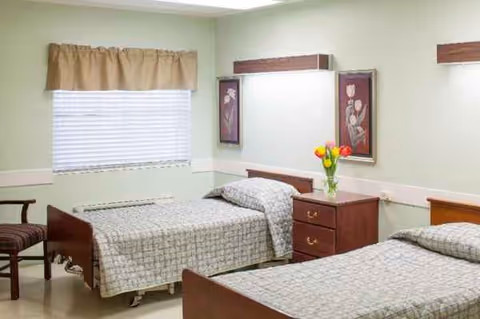 Shared bedroom in a senior care facility with two beds, dressers, a window, and green walls.
