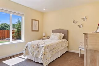 A bright bedroom with a single bed covered in a floral bedspread, a window showing a view of a fenced garden with trees, a framed picture on the wall, butterfly wall decorations, a wooden dresser, and a chair.