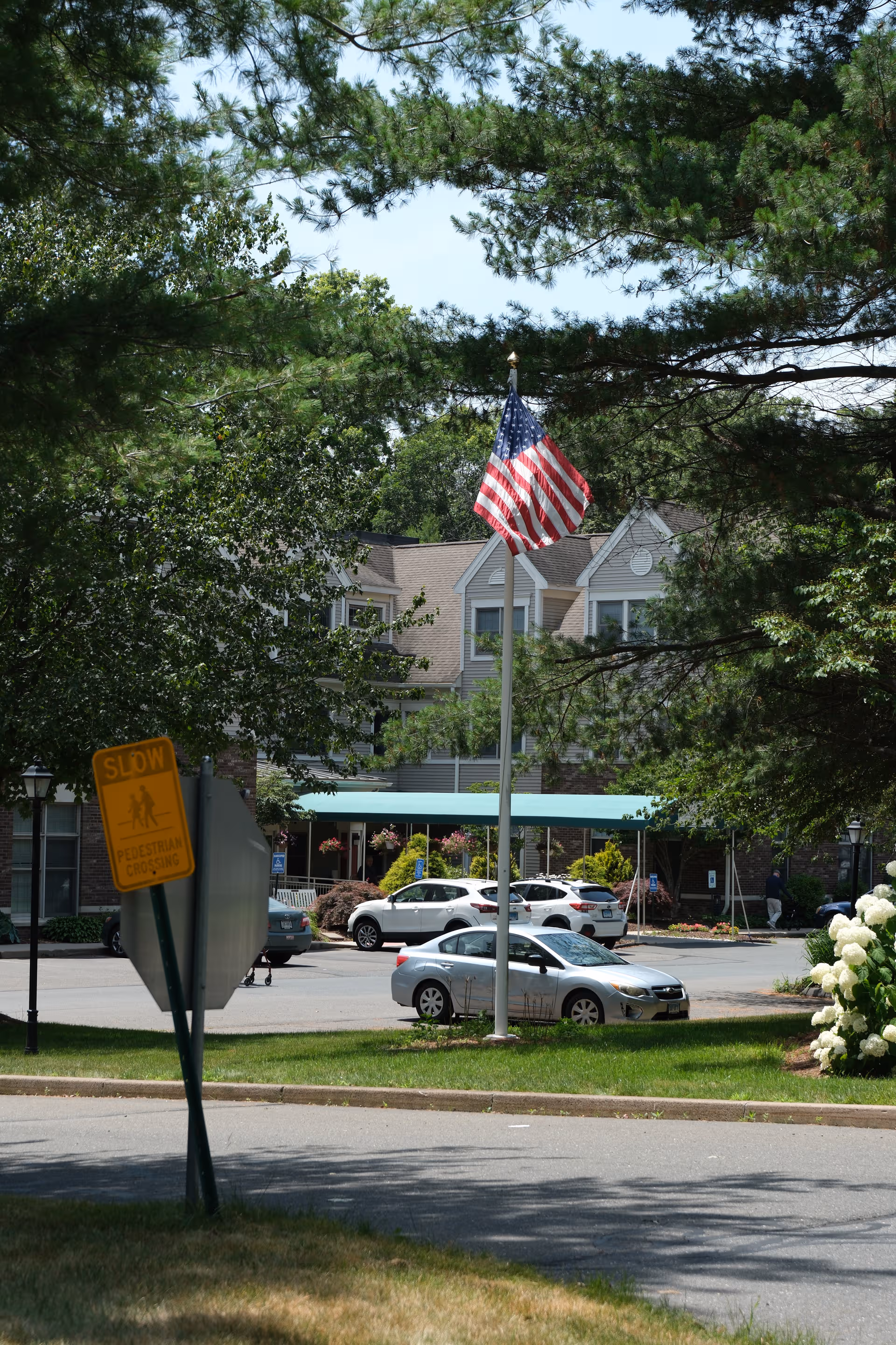 Exterior of a senior living facility with trees, a waving American flag, and parked cars.