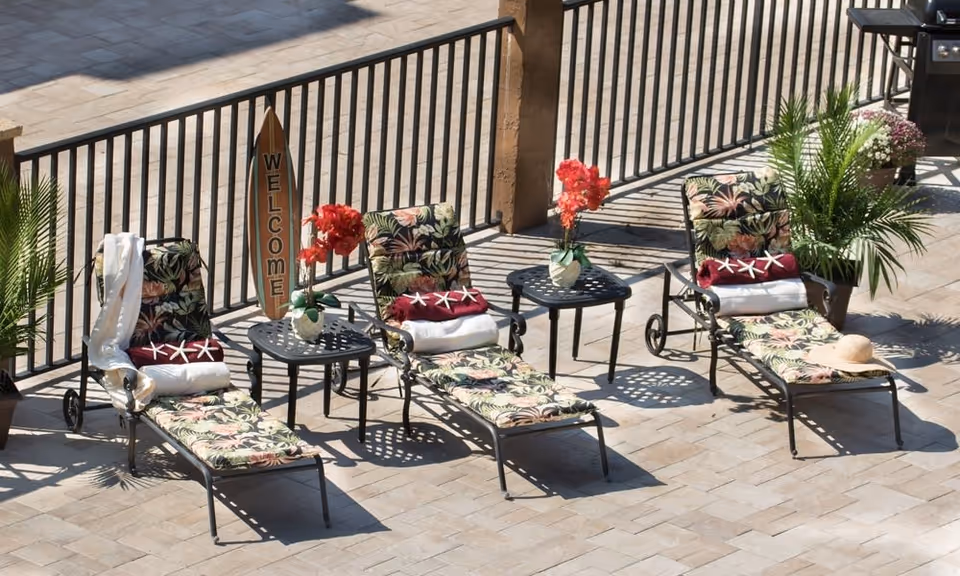 Three floral-patterned lounge chairs with cushions and towels arranged on a tiled patio next to a black metal railing. Two small black tables with flower vases and red flowers are placed between the chairs. A wooden surfboard-shaped sign with the word 'WELCOME' is positioned behind the middle chair. Potted green plants are placed on either side of the seating area.