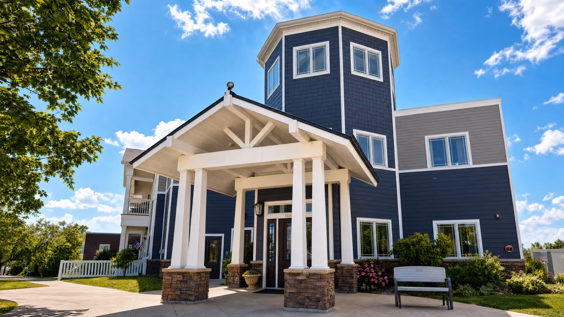 Blue multi-story senior living building with white entry columns and a bench outside.