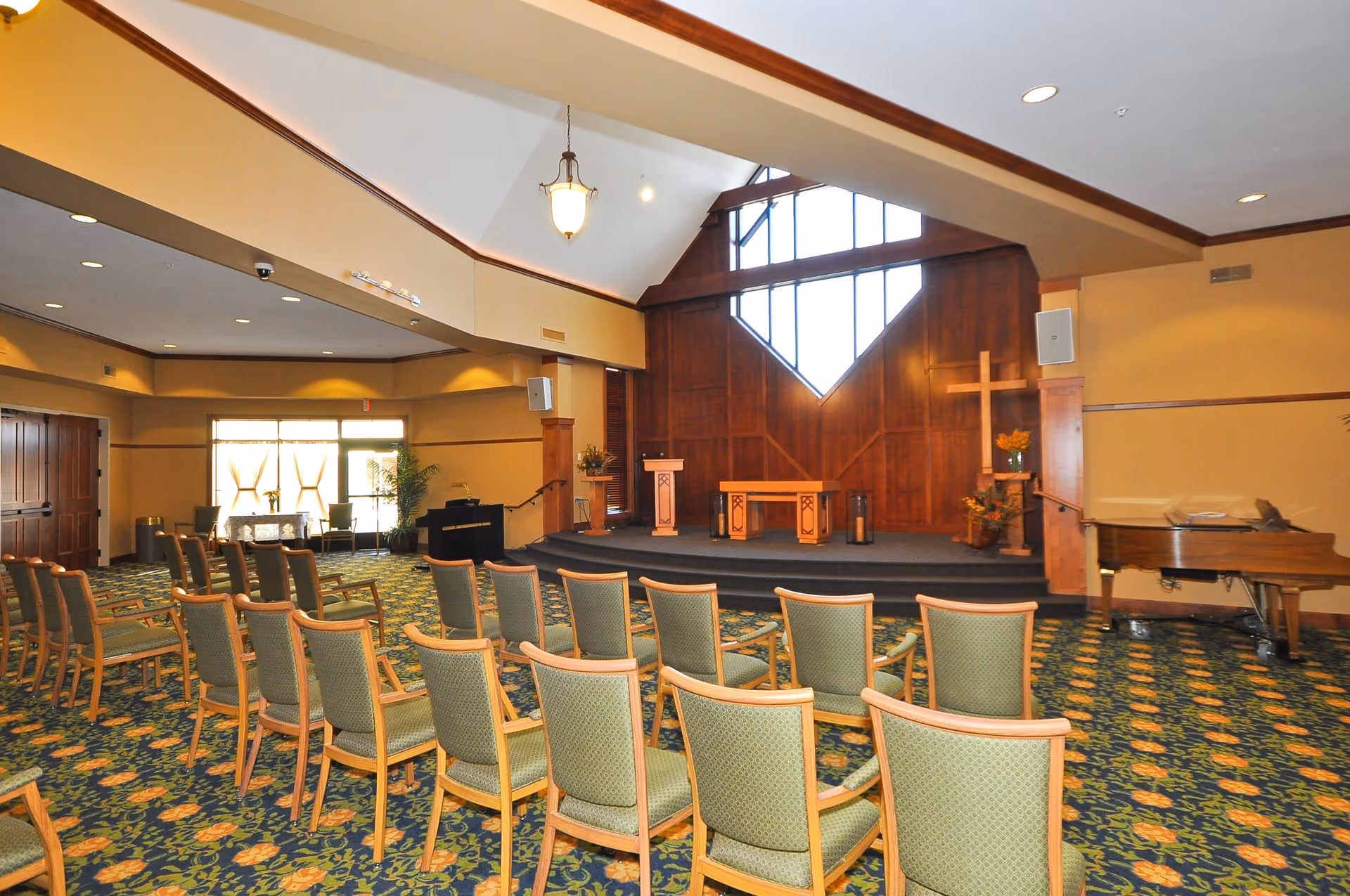Large chapel-style multipurpose room with rows of chairs facing a raised wooden stage with a lectern, cross, and piano.
