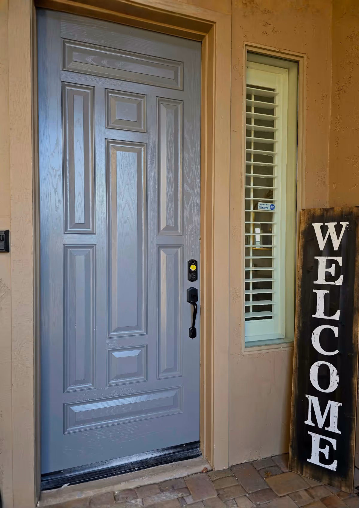 Front entrance with a grey paneled door, window, and a tall 'WELCOME' sign outside.