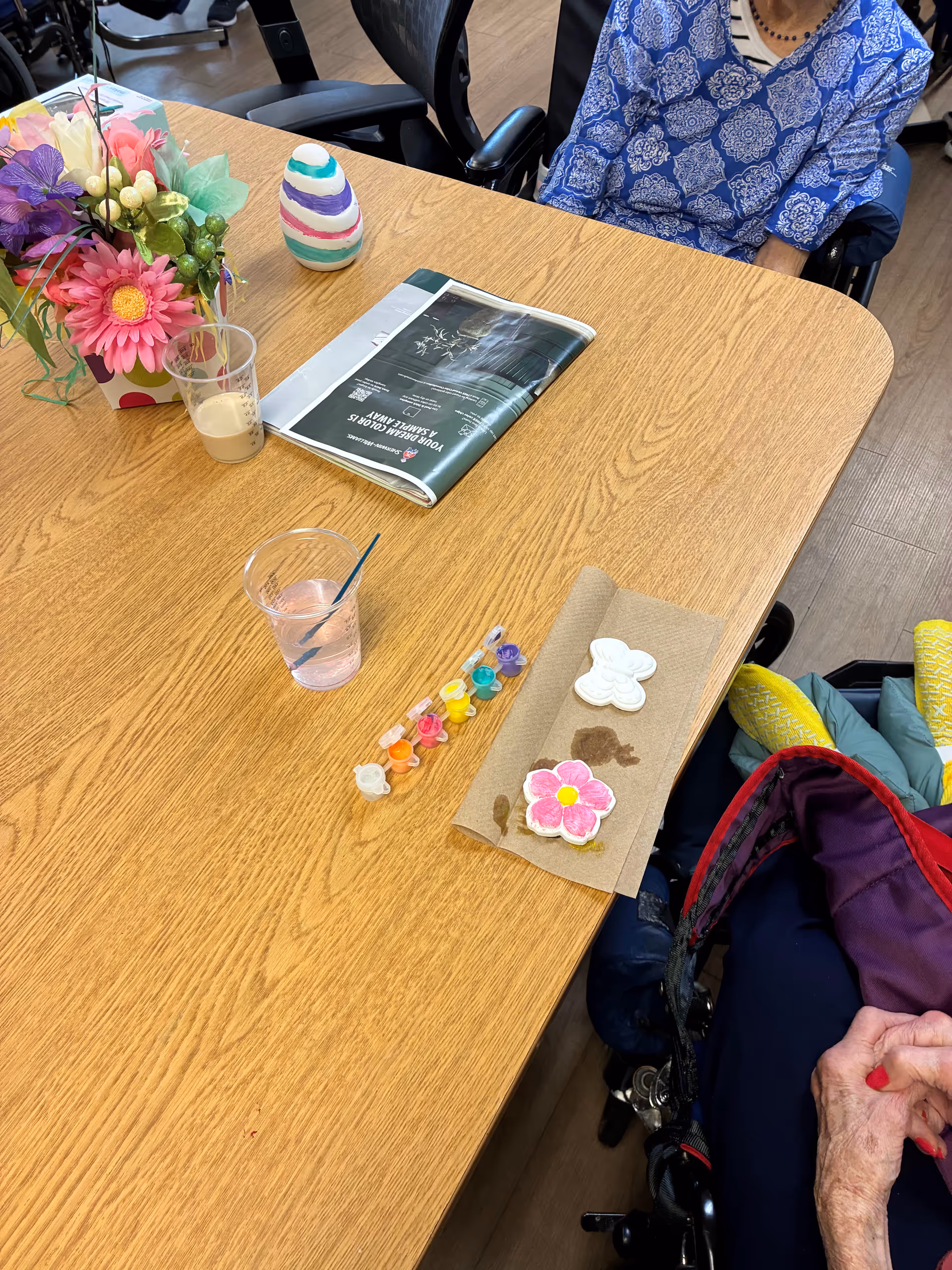 Senior resident in a wheelchair sits at a wooden table with crafts, flowers, and a magazine.