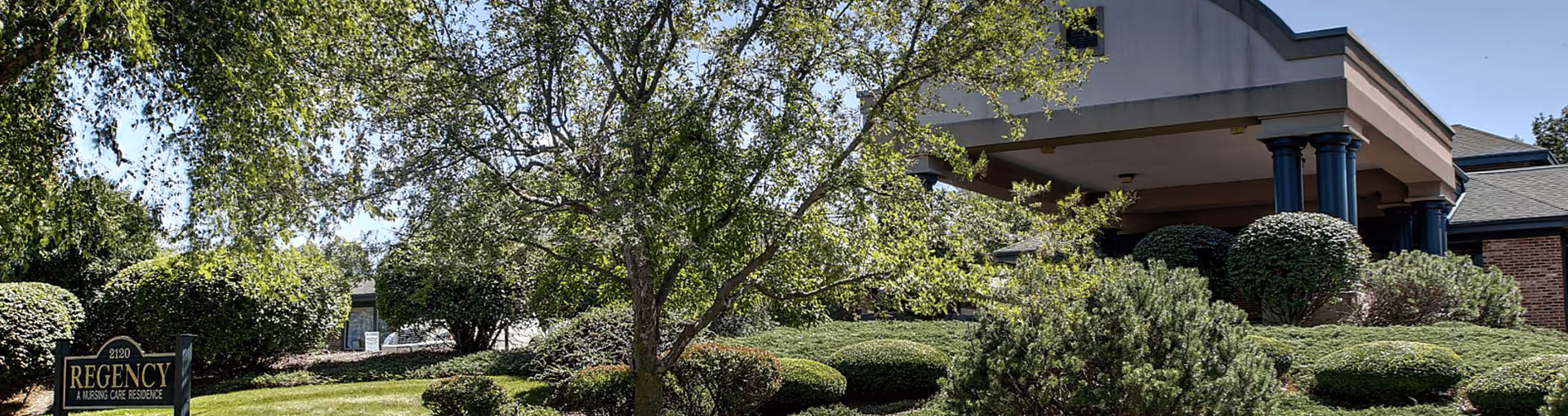 Exterior front of the Regency nursing care residence with landscaped shrubs, trees, and an entrance canopy.