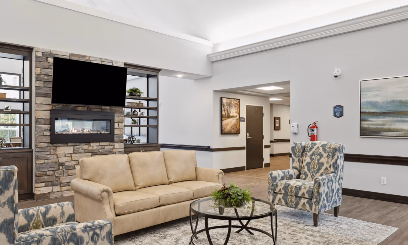 A furnished common living area with a beige sofa and patterned armchairs facing a stone fireplace and wall-mounted TV.