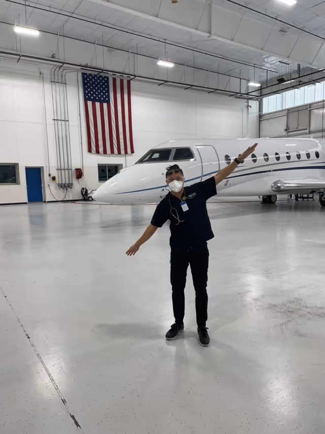 A person wearing a mask stands inside a hangar beside a private jet.