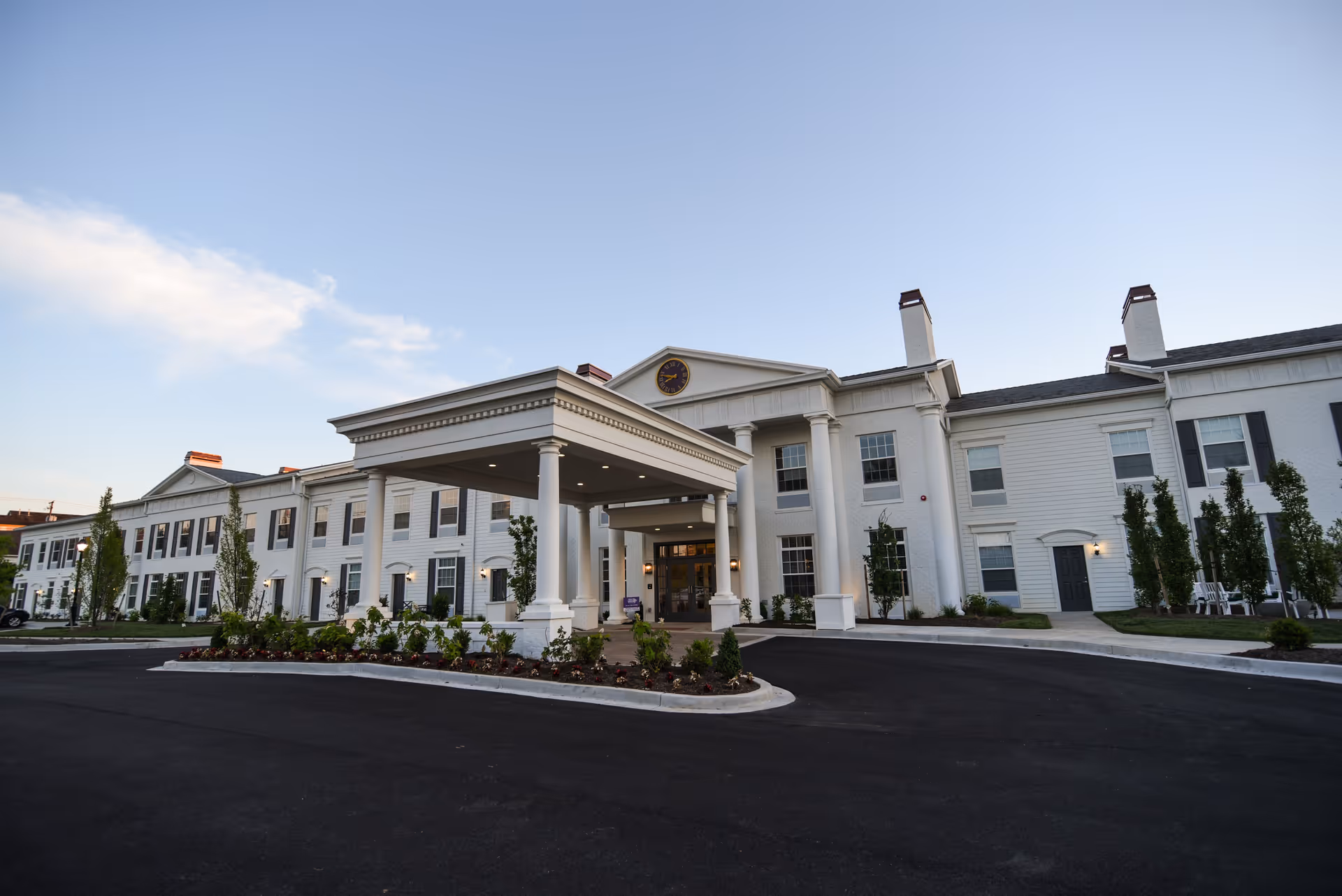 Exterior view of a large, white senior living facility building with a covered entrance supported by columns, surrounded by landscaped greenery and a paved driveway under a clear sky.