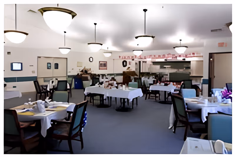 A dining room in an assisted living facility with multiple tables covered with white tablecloths, set with plates, cups, and napkins. The room has several chairs around each table, ceiling lights, and a carpeted floor. The walls are light-colored with some decorations and a clock.