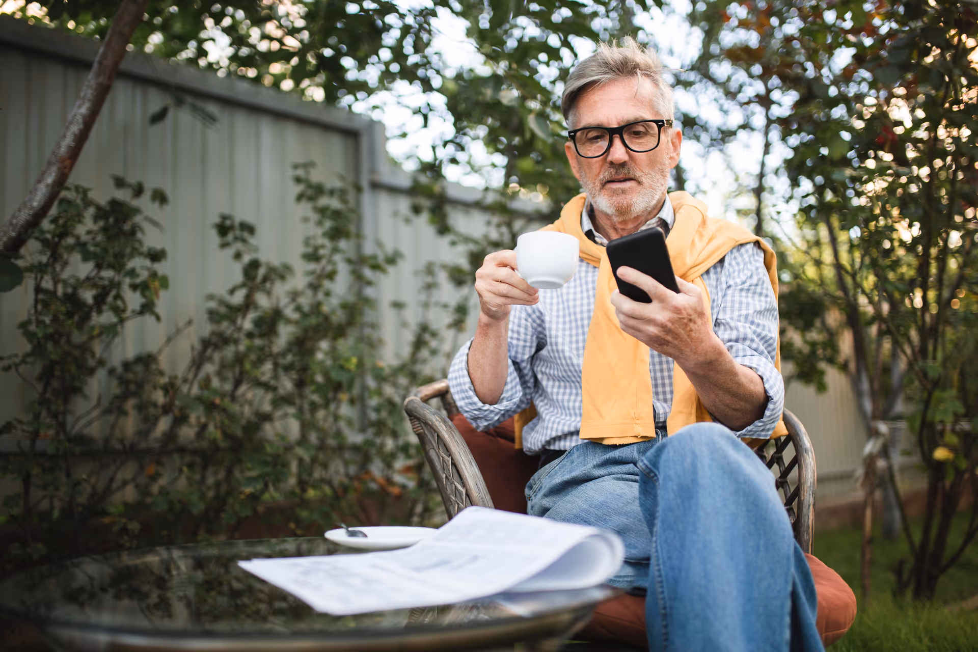 Older man sits outdoors at a garden table, holding a mug and smartphone.