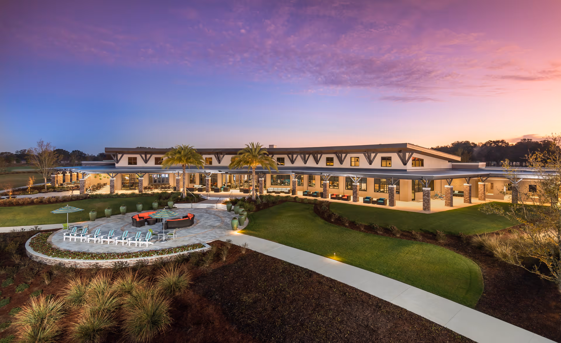 Wide exterior view of a modern resort community building at sunset with a landscaped garden, palm trees, outdoor seating area with lounge chairs and umbrellas, and a paved walkway.