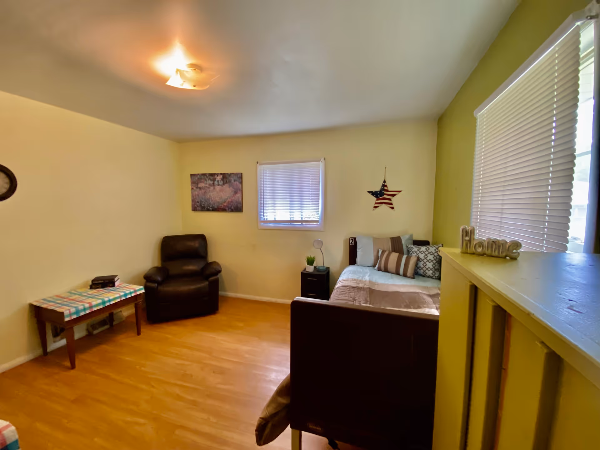 Cozy living room with a recliner, bed, side table, large windows, and a decorative 'Home' sign.