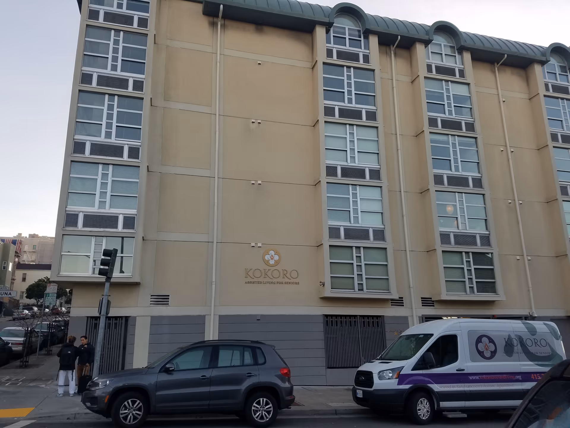 A multistory beige Kokoro Assisted Living building exterior with windows and parked vehicles.