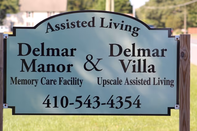 A light blue sign reading Assisted Living Delmar Manor & Delmar Villa with a phone number.