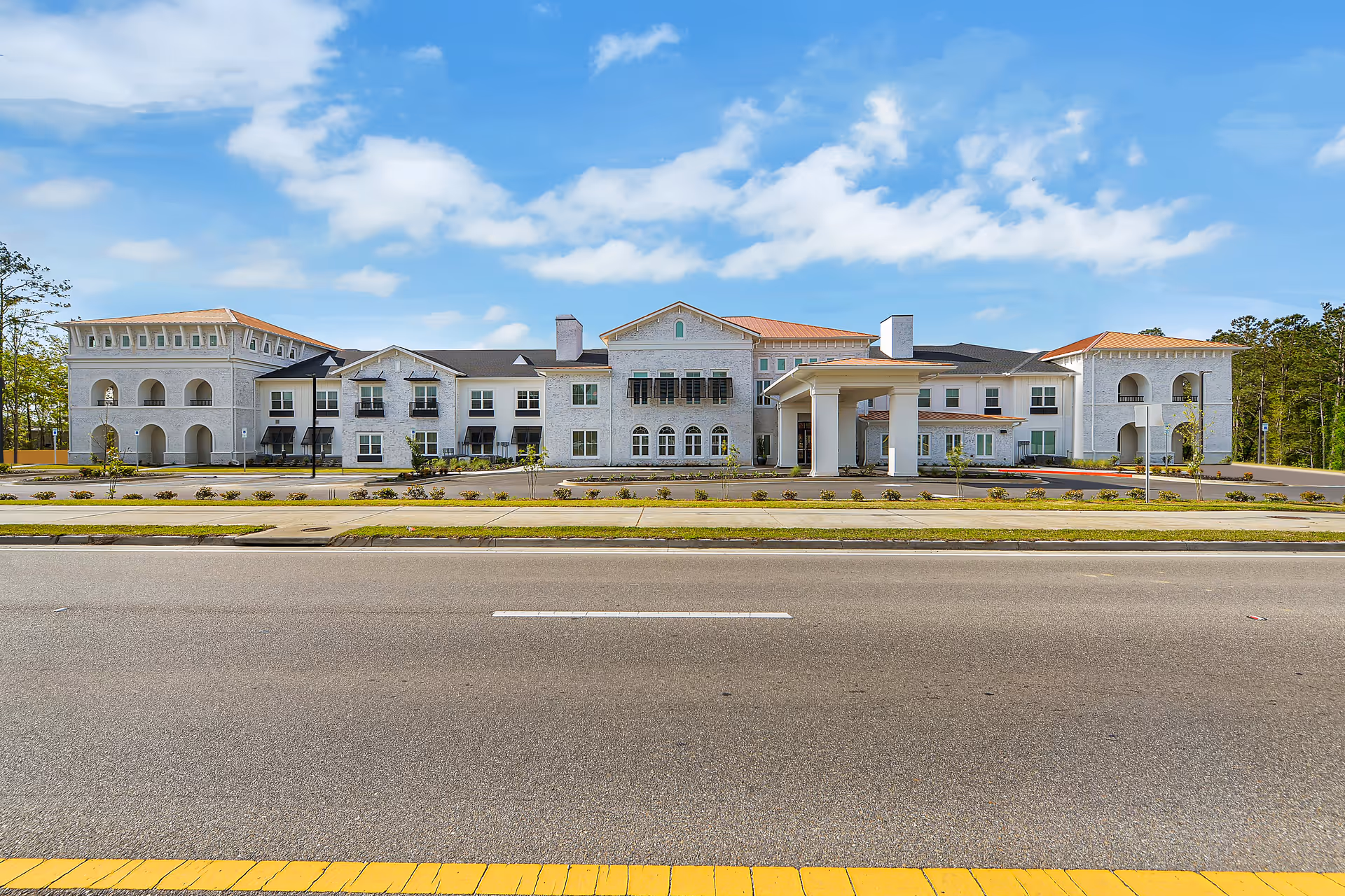 Front exterior view of a large, two-story senior living facility building with white walls, multiple windows, and a covered entrance. The building is set against a blue sky with scattered clouds and is surrounded by a paved road and landscaped greenery.