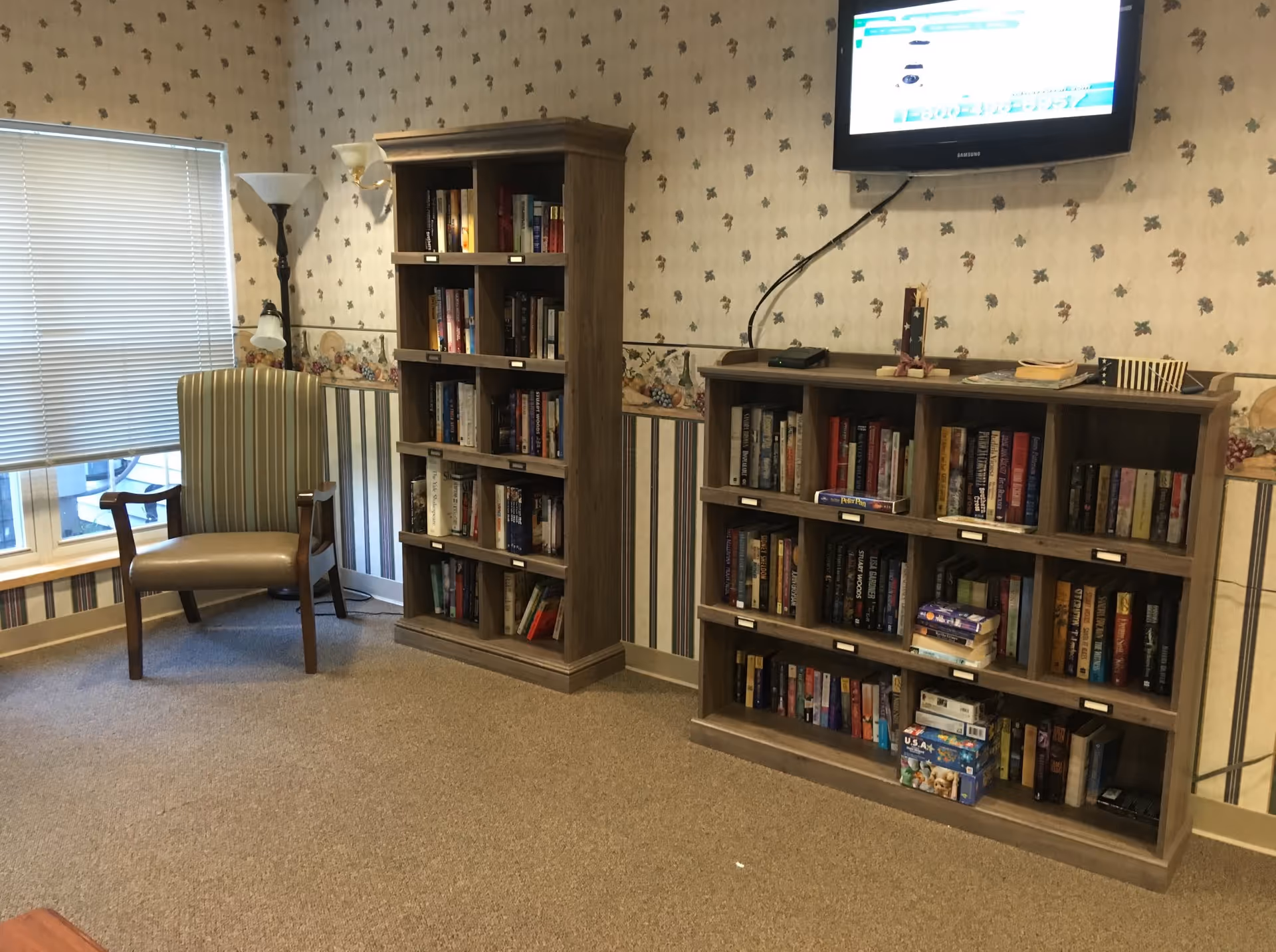 A cozy reading area with tall bookshelves, a chair, window blinds, and a wall-mounted TV.