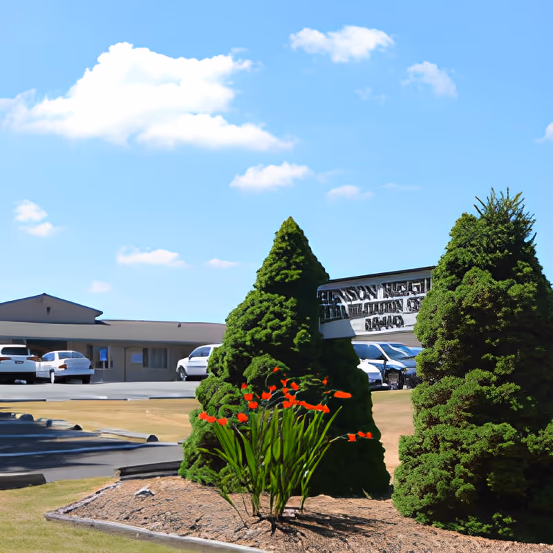 Outdoor view of Benson Heights Rehabilitation Center showing a parking lot with several cars, manicured bushes, and a flower bed with red flowers under a blue sky with scattered clouds.