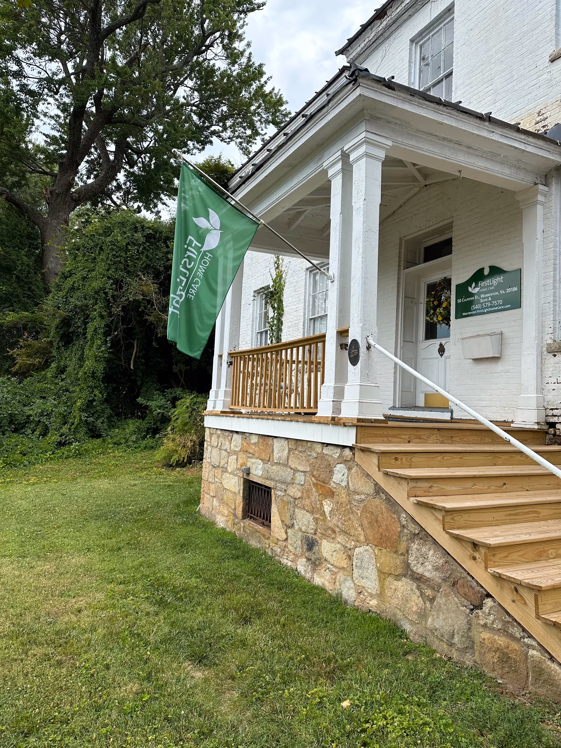 White brick building with a wooden porch, stone foundation, and a green flag banner.