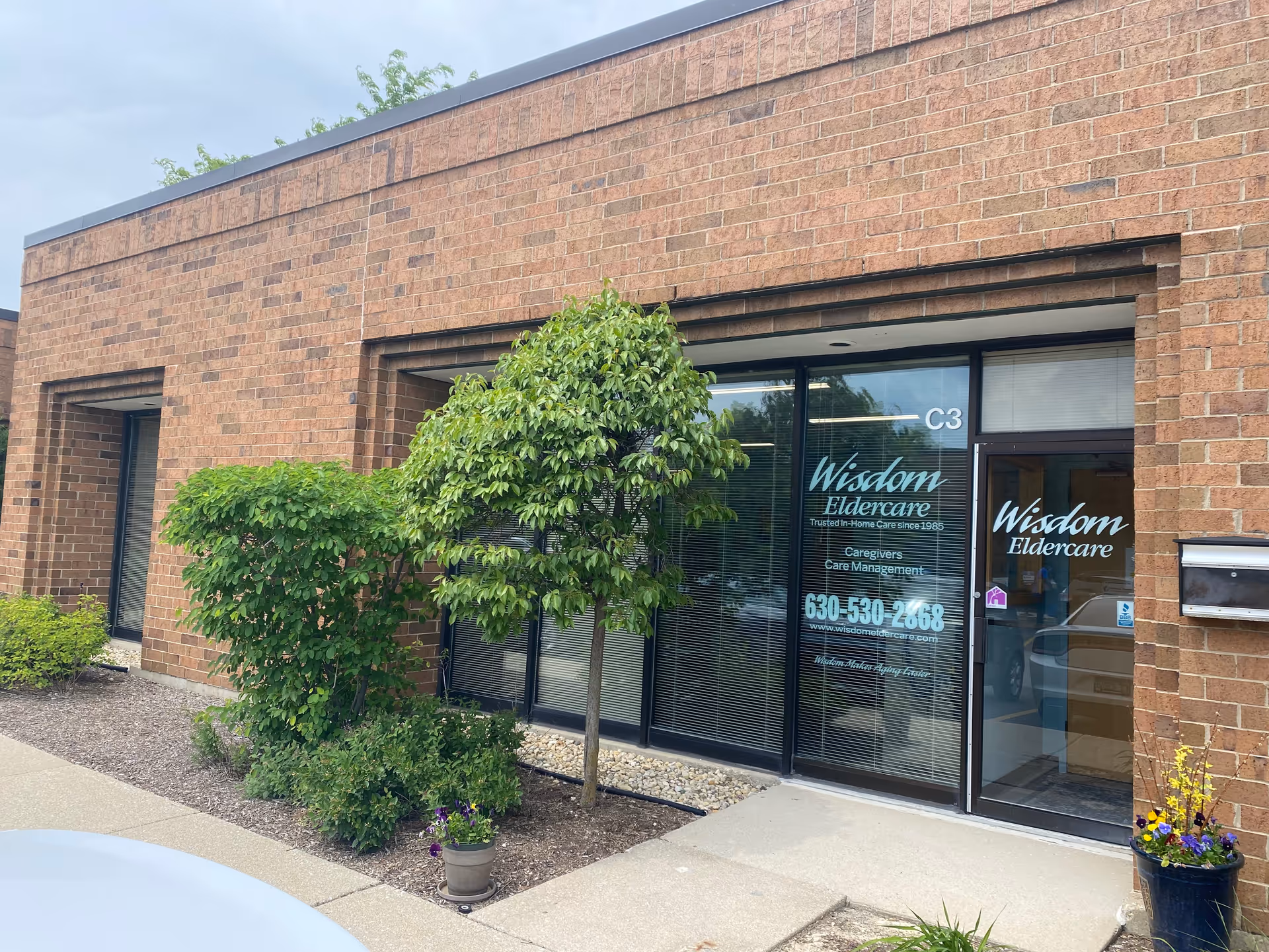 Exterior view of Wisdom Eldercare entrance with brick building, glass doors, and surrounding shrubs.