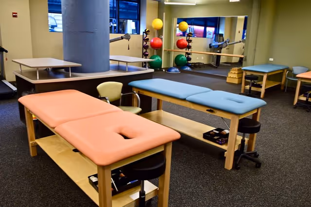 Bright physical therapy room with padded treatment tables, chairs, and colorful exercise balls.