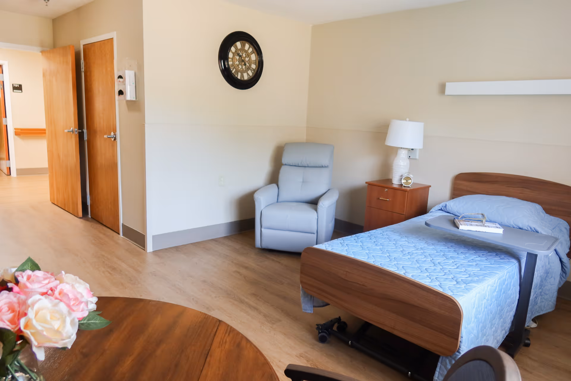 A senior living facility patient room with a bed, blue bedding, chair, and nightstand.