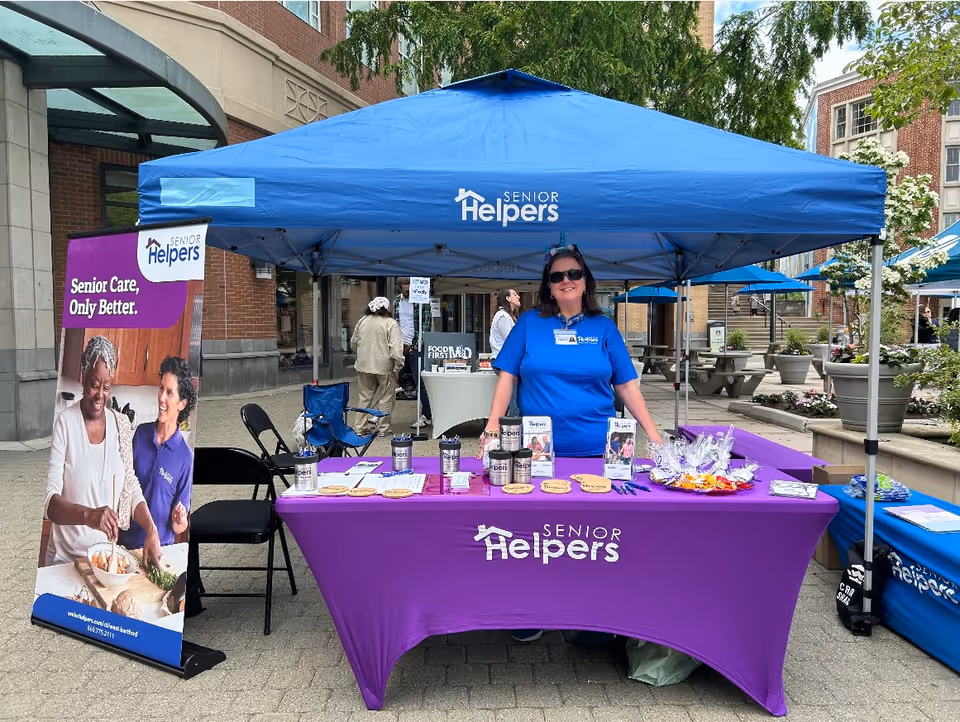 Outdoor Senior Helpers booth with purple tablecloth, blue canopy, and staff member.