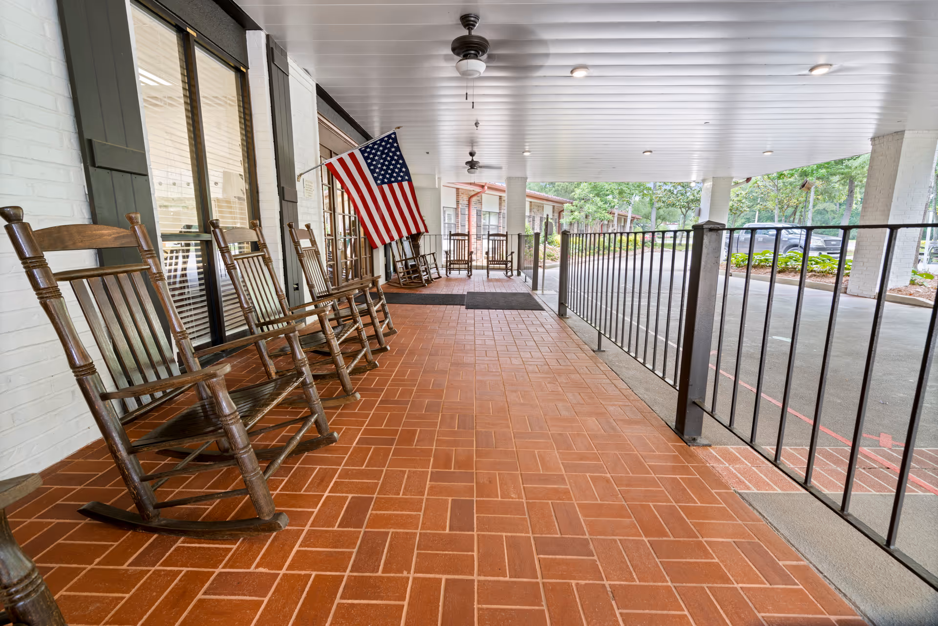 Covered outdoor porch area with several wooden rocking chairs lined up along a white brick wall. An American flag is mounted on the wall near the chairs. The porch has a tiled floor and ceiling fans, with a black metal railing separating the porch from the driveway and greenery outside.