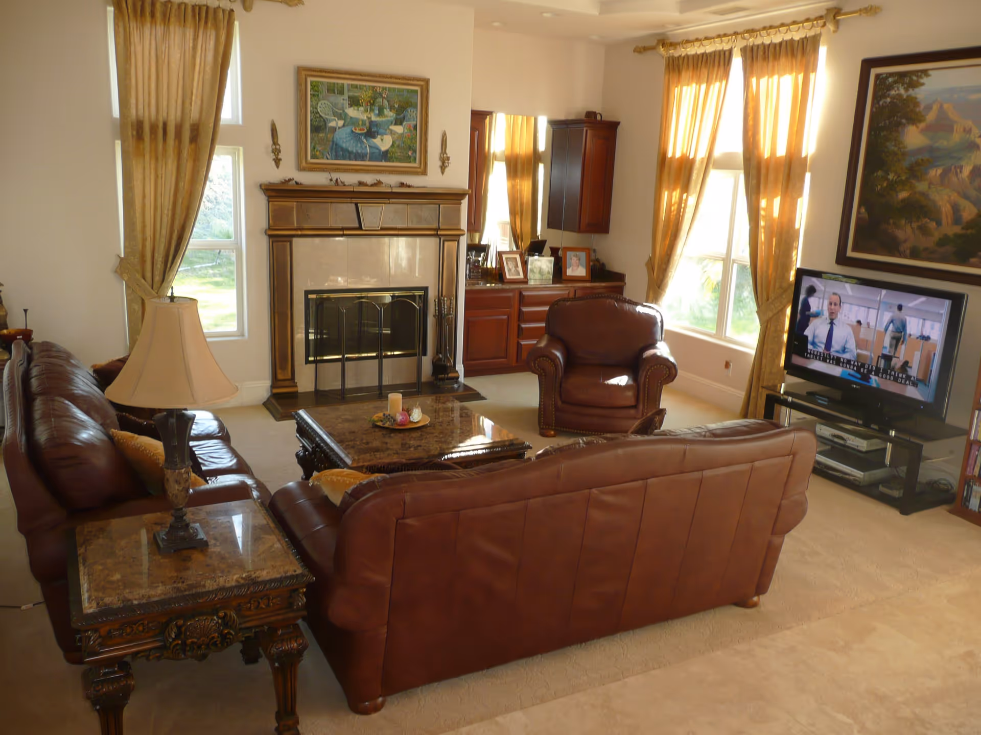 A cozy living room with brown leather sofas and an armchair arranged around a marble coffee table. There is a fireplace with a decorative screen and a painting above it. Large windows with golden curtains let in natural light. A flat-screen TV is on a stand showing a man in an office setting. A side table with a lamp and a wooden cabinet with framed photos are also visible.