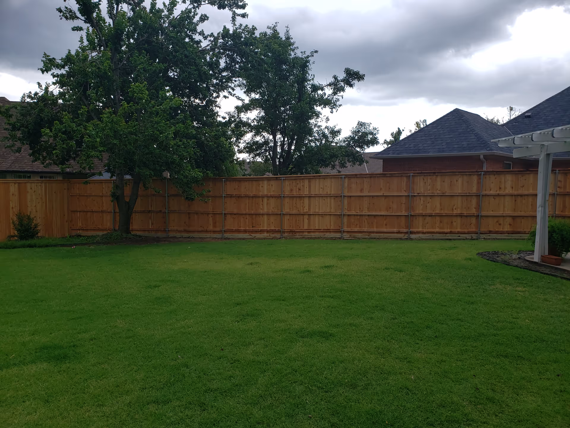 Backyard lawn with a wooden privacy fence, trees, and a corner patio.