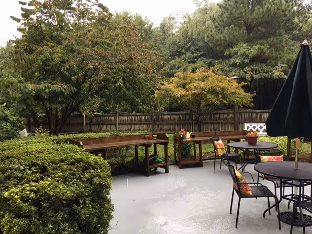 Outdoor patio area with round tables and chairs, some with colorful cushions, and a large closed umbrella. There are wooden benches along the fence, potted plants on the tables and benches, and lush green trees and bushes surrounding the space.