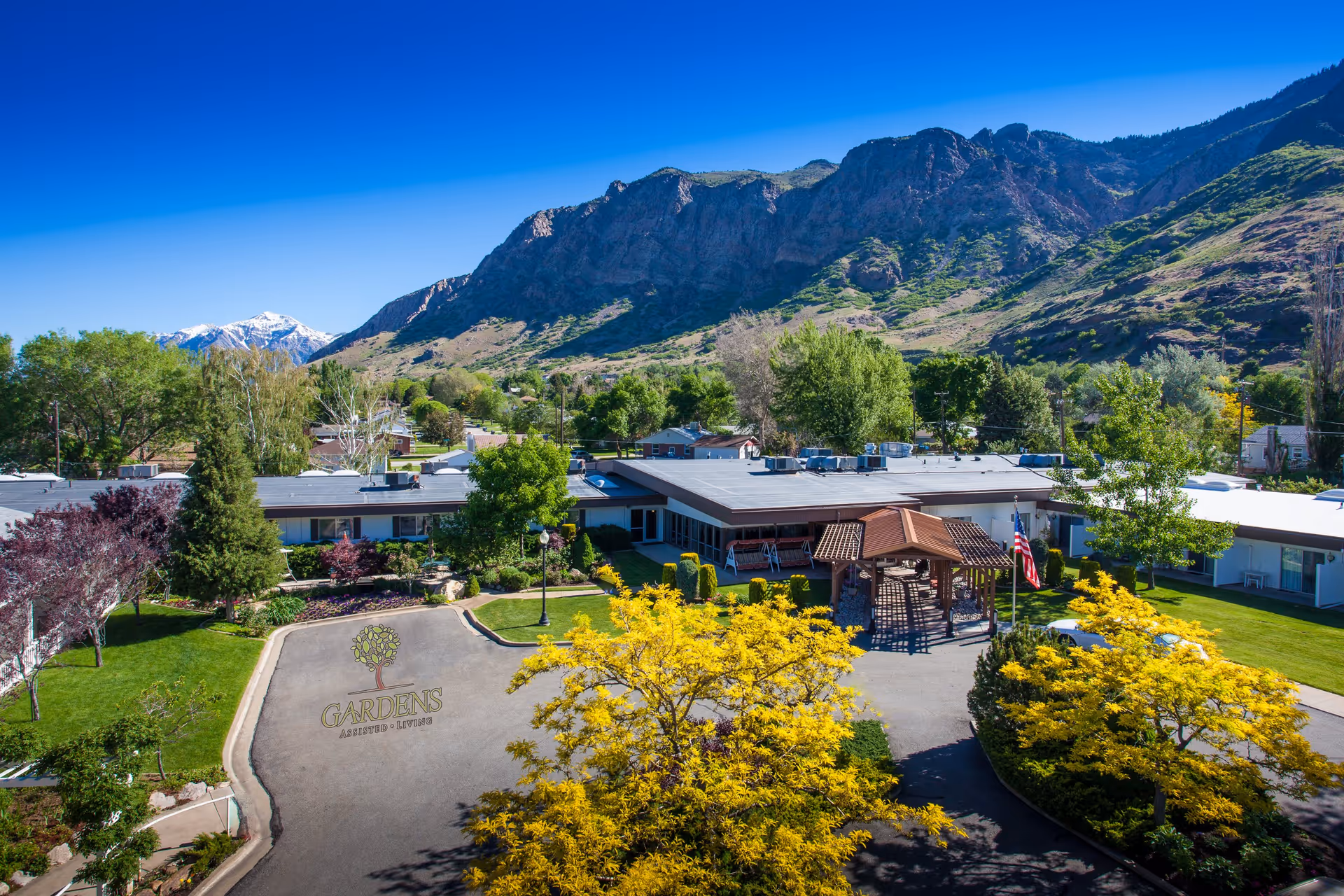 Aerial view of the Gardens Assisted Living complex with a central covered entrance, landscaped grounds and mountains in the background.
