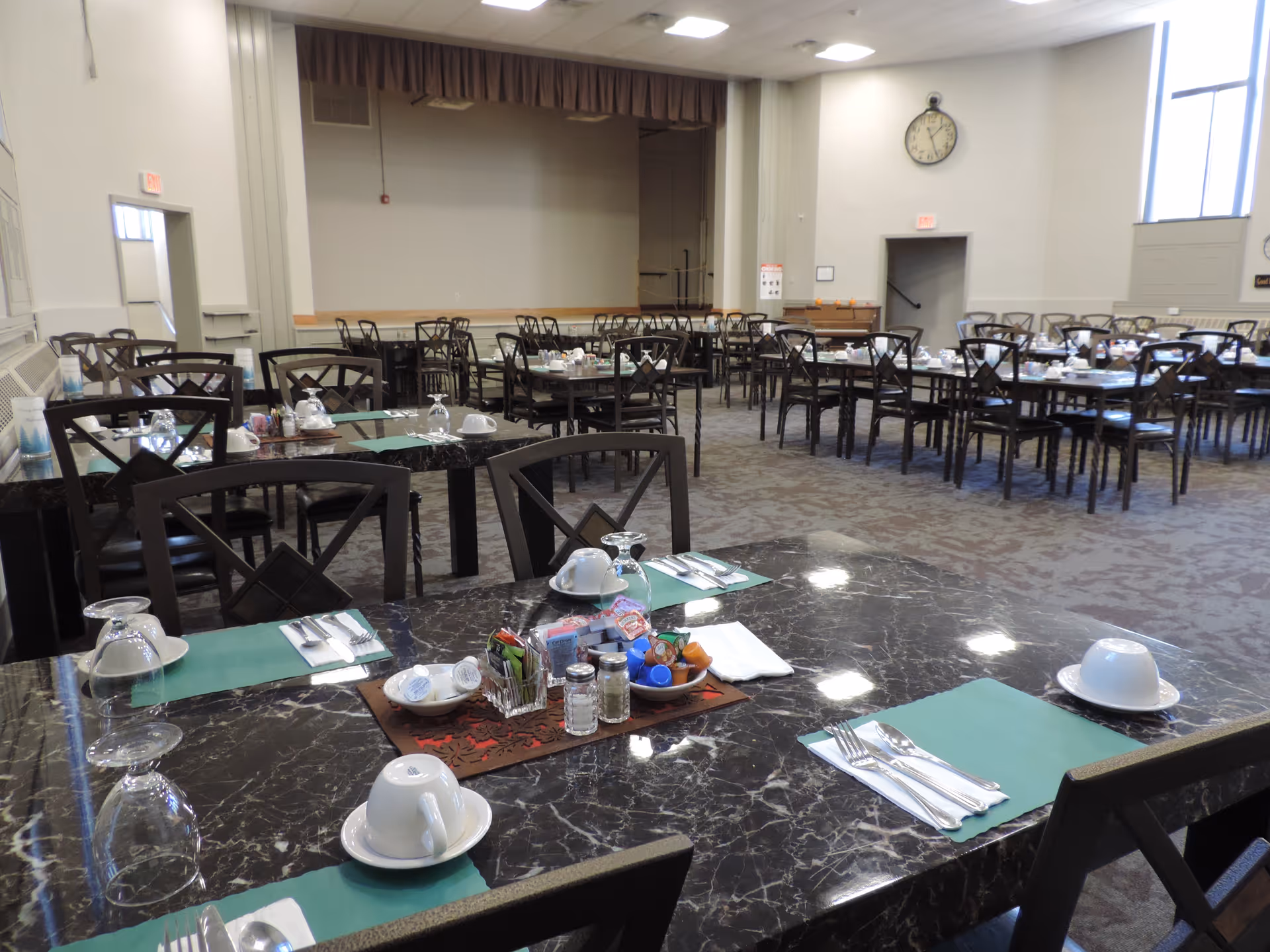 A spacious dining room with multiple dark marble tables set with green placemats, white cups and saucers, silverware, and glassware. The room has many wooden chairs with cross-back designs, a carpeted floor, large windows letting in natural light, and a clock on the wall. There is a small stage area at the far end of the room.
