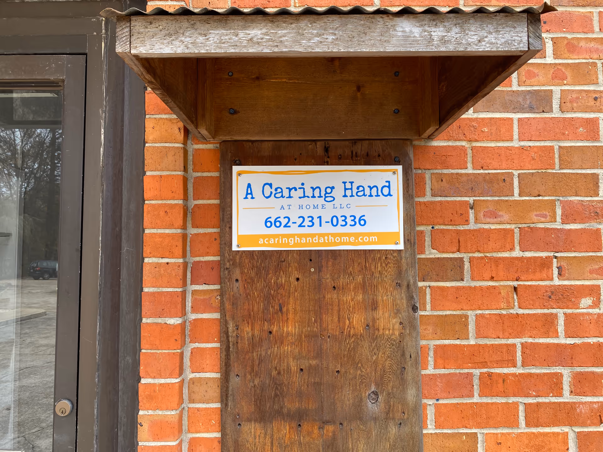 Brick exterior with a wooden sign reading A Caring Hand AT HOME LLC beside a door.