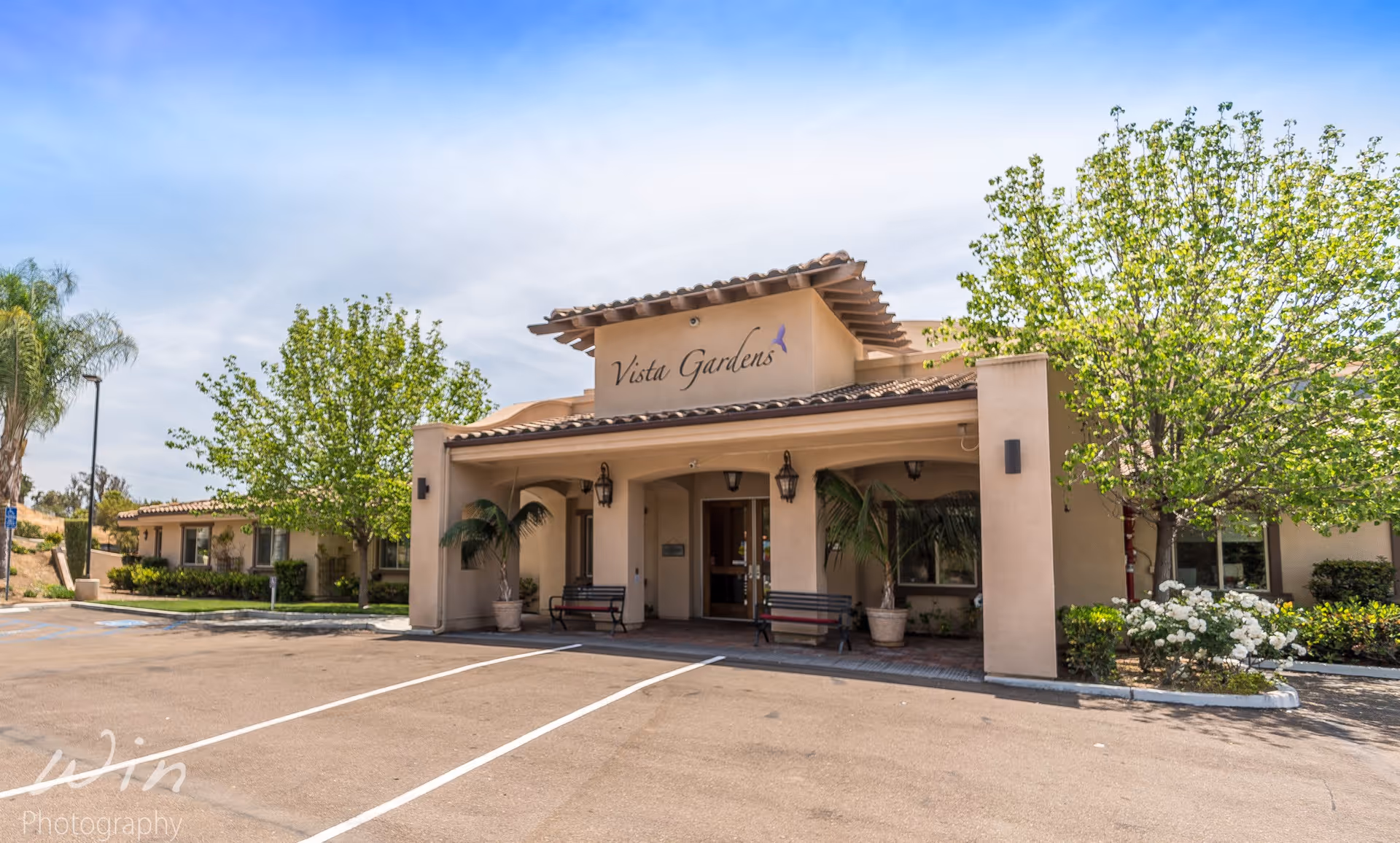 Exterior view of Vista Gardens Memory Care facility showing the entrance with a covered porch, two benches, potted plants, and surrounding greenery including trees and bushes under a partly cloudy sky.
