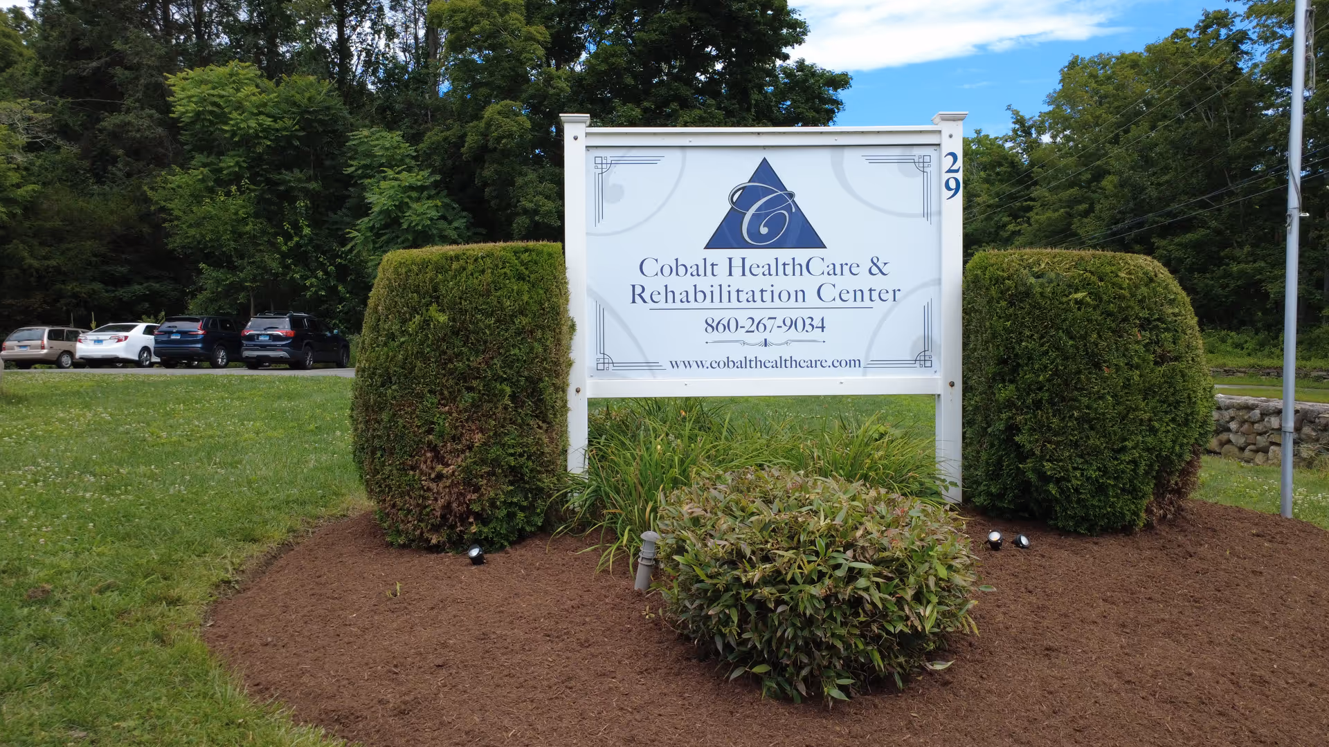 Exterior view of a sign for Cobalt HealthCare & Rehabilitation Center with shrubs and parked cars.