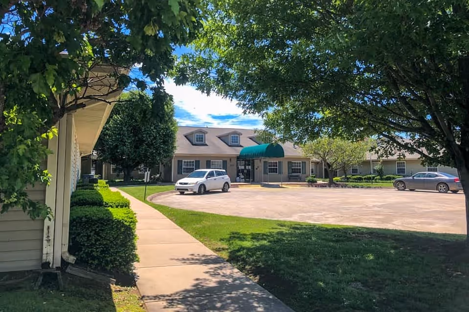 Exterior view of Peachtree Village Assisted Living of Farmington showing a single-story building with a green awning over the entrance, a parking lot with two cars, and a sidewalk surrounded by green grass and trees under a blue sky with some clouds.