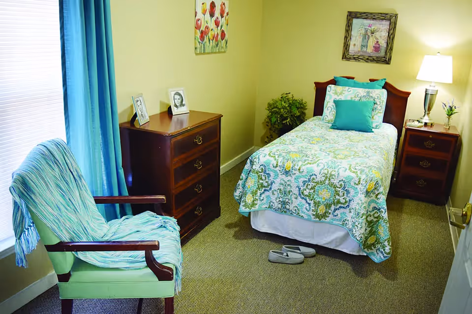 A cozy bedroom with a single bed, dresser, chair, one nightstand, and slippers.