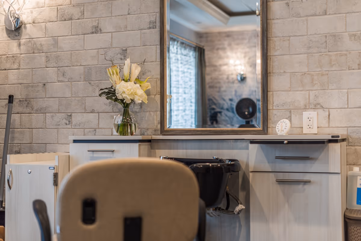 Interior salon-style grooming station with a mirror, hair-washing sink, chair, and a vase of flowers against a light brick wall.