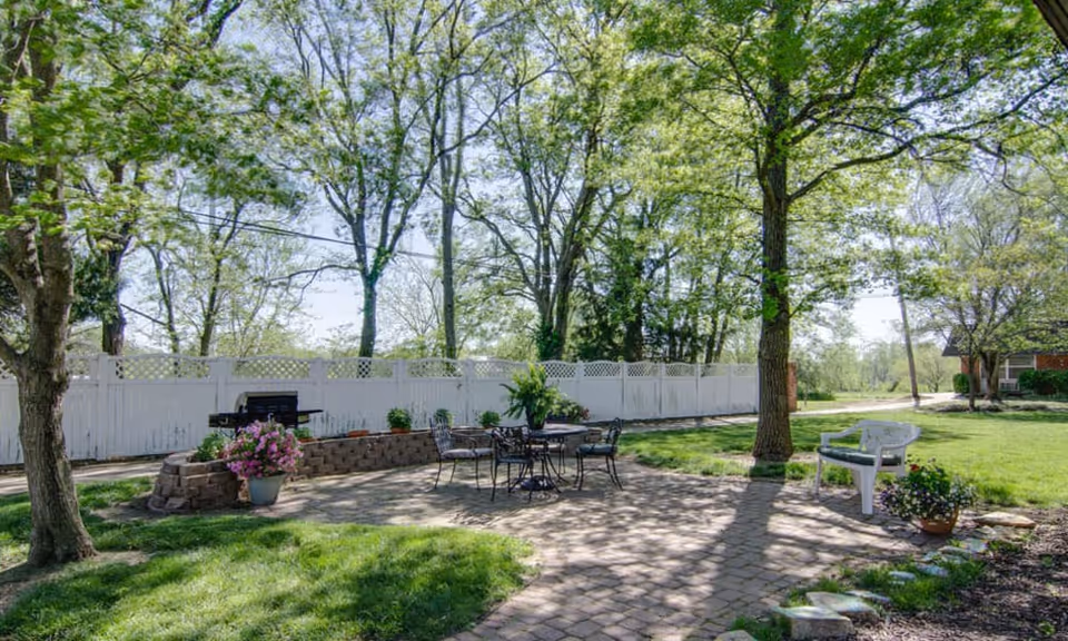 Sunny outdoor patio with a metal table and chairs, a grill, potted flowers, trees and a white privacy fence.
