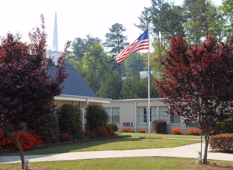 Front exterior of a single-story assisted living building with an American flag on a flagpole, landscaped lawn and trees.