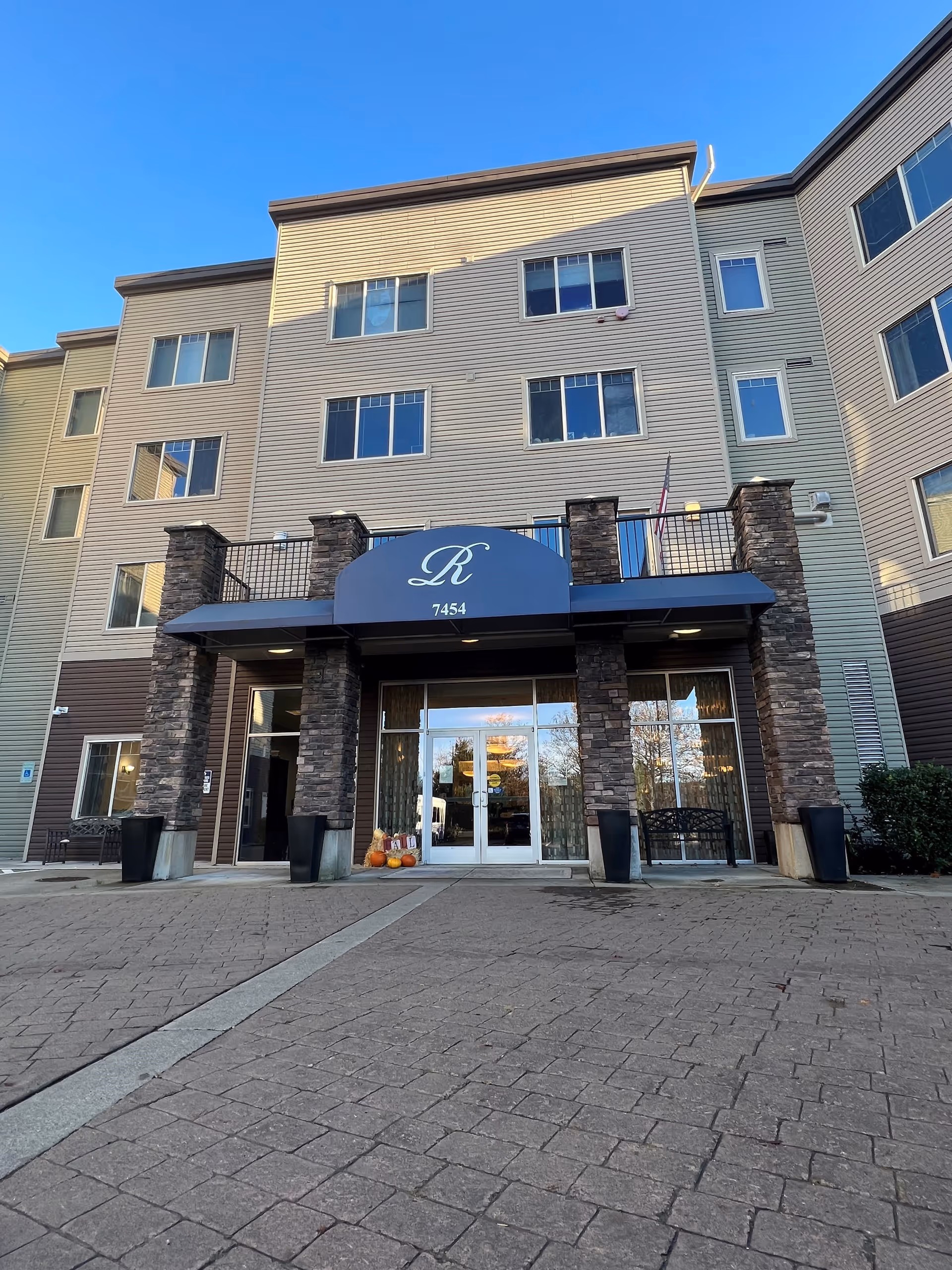 Front entrance of a multi-story residential building with a blue awning marked 'R' and '7454', stone pillars, glass double doors and pumpkins on the doorstep.
