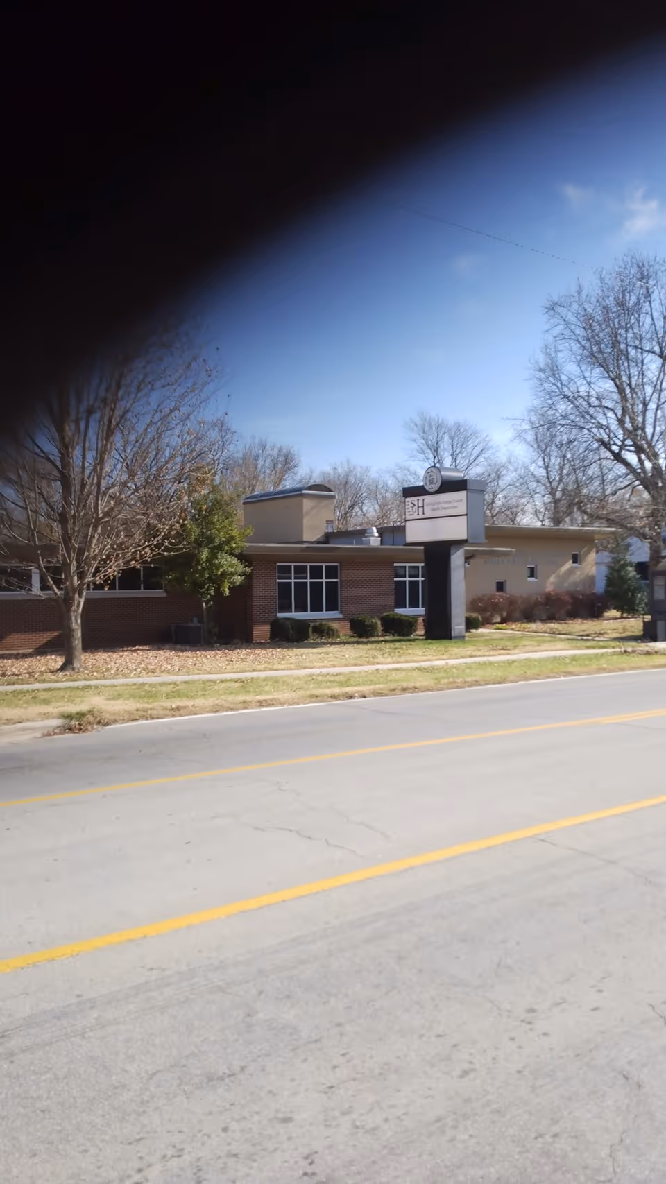 Exterior view of a brick public health center with a sign, trees, and a street.
