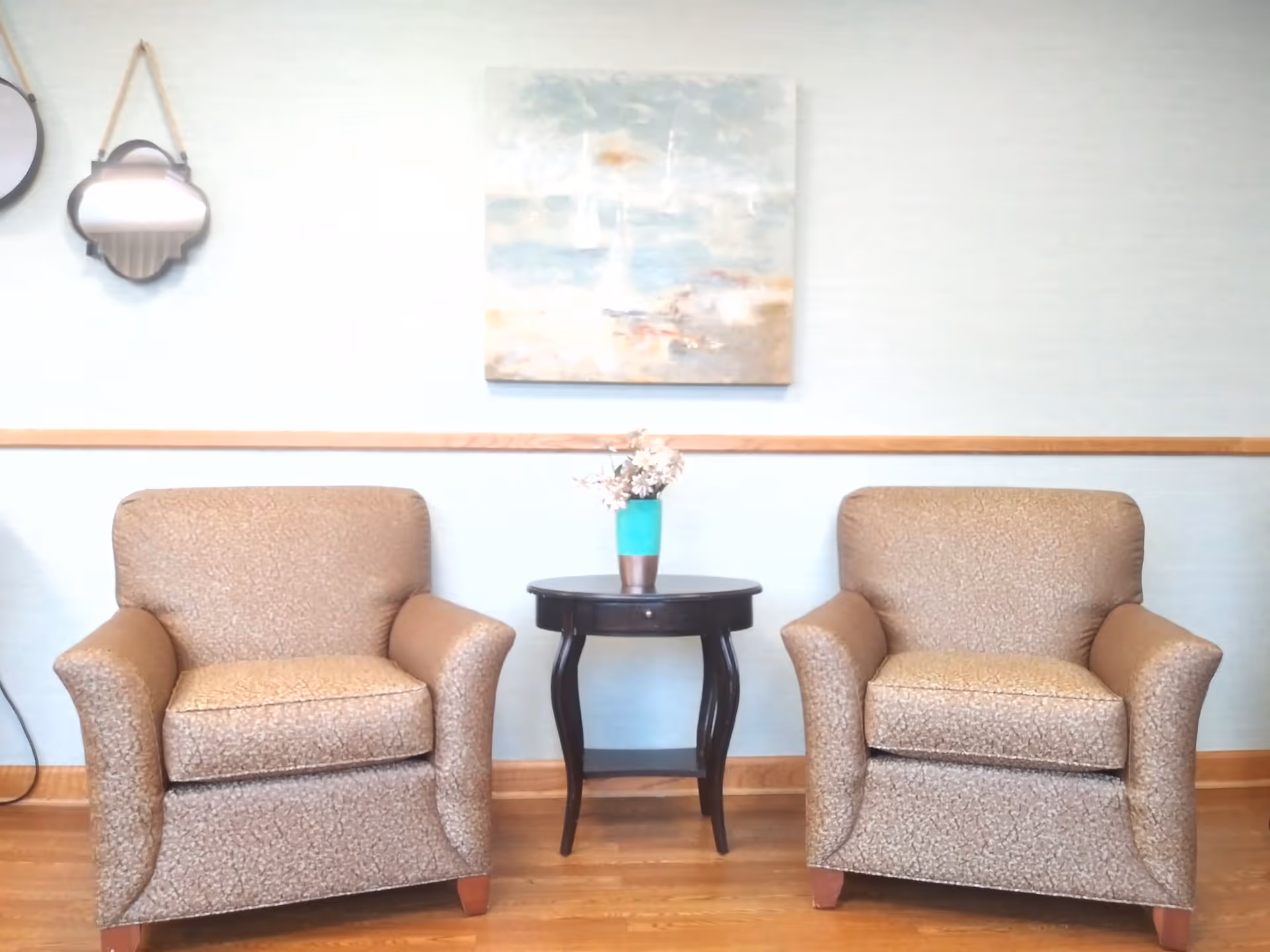 Two beige armchairs flank a small round table with a turquoise vase in a sunny lounge.